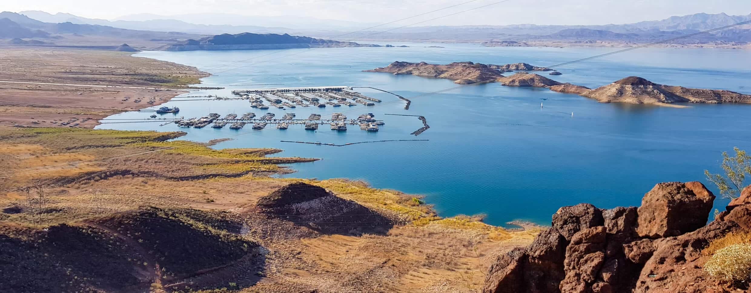 An aerial view shows a wide blue lake with many boats docked in a marina, surrounded by dry, rocky land.
