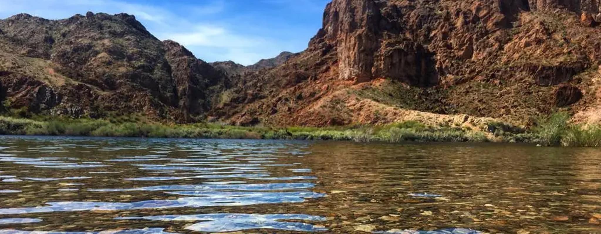 A wide river with ripples reflects a blue sky, flowing past a green and brown rocky mountain landscape.
