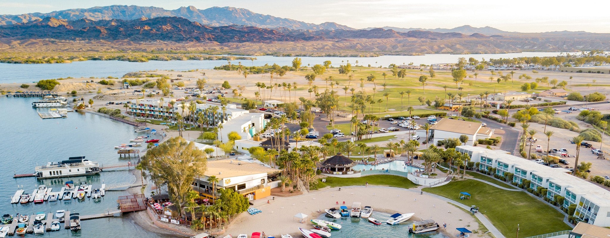 An aerial view of a resort town with a marina, hotels, palm trees, and mountains in the distance.