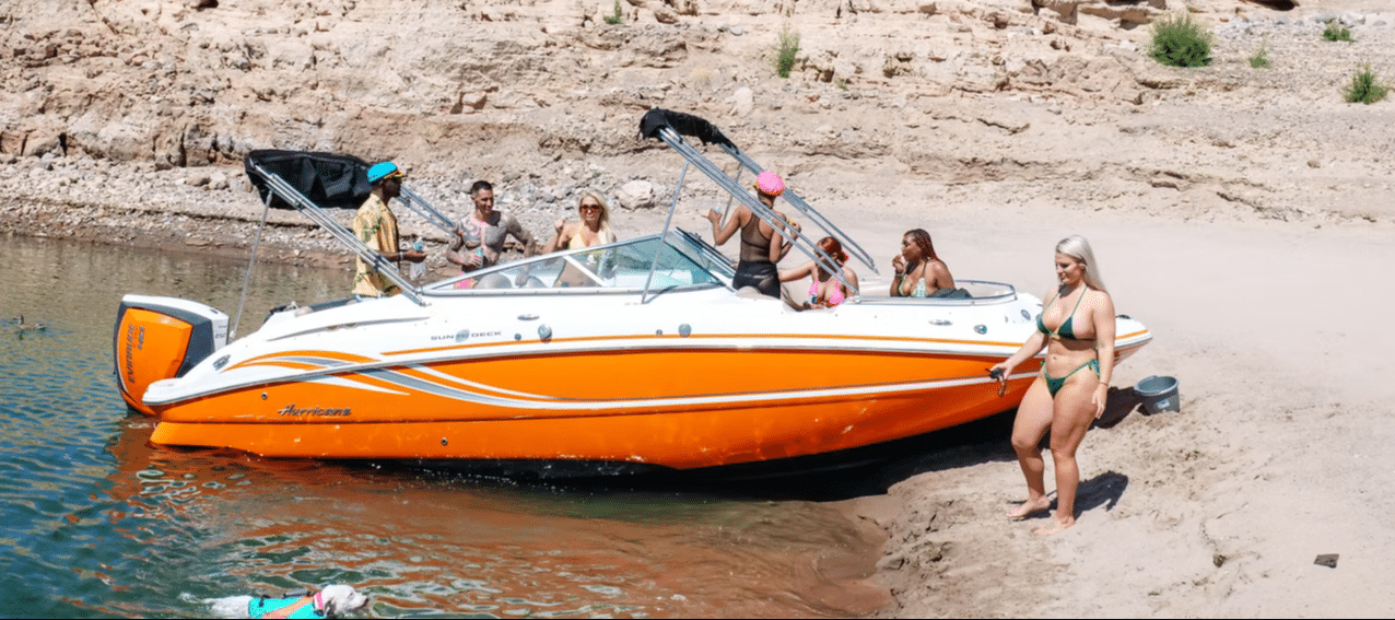 A group of people in swimsuits are on and around an orange and white "Hurricane" boat at a sandy beach.