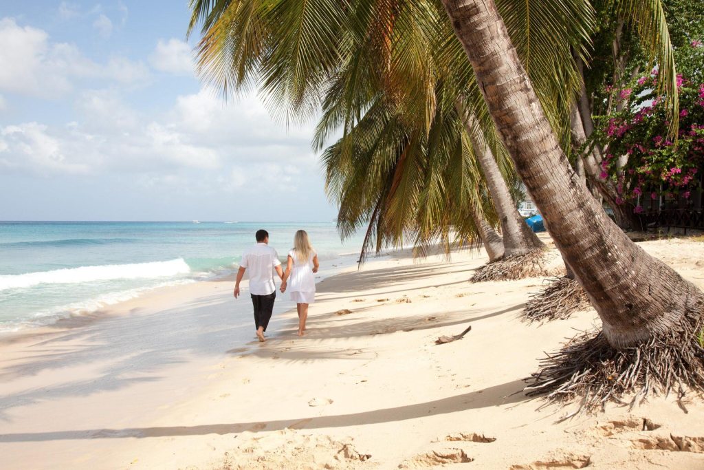 A man and woman in white walk barefoot, holding hands on a sandy beach under palm trees.