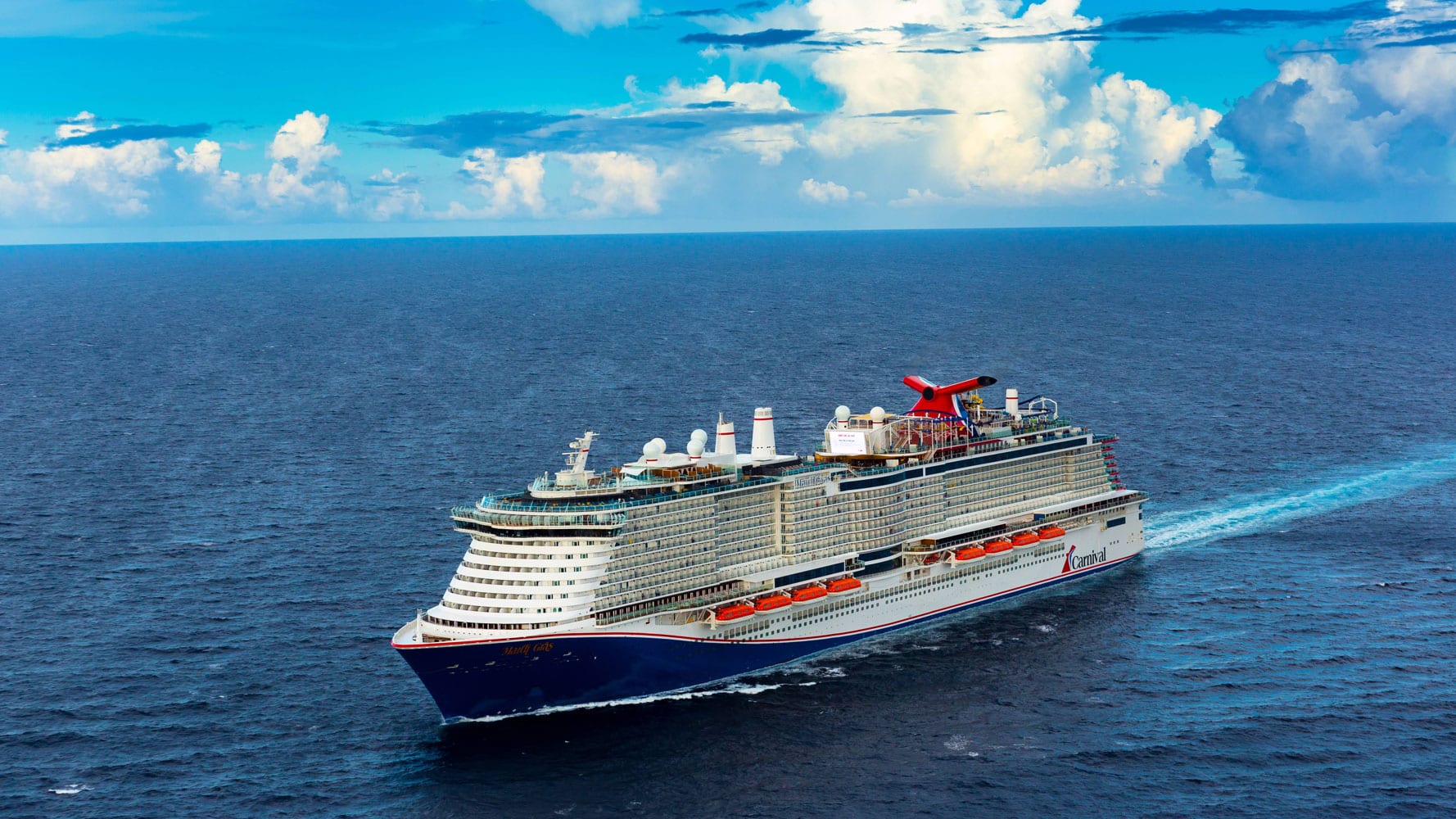 A large "Carnival" cruise ship with a red funnel sails on blue ocean water under a partly cloudy sky.