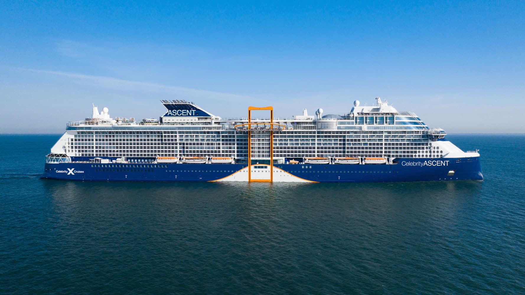 A large blue and white cruise ship named "Celebrity ASCENT" with a prominent orange structure on its side floats on calm blue water under a clear sky.