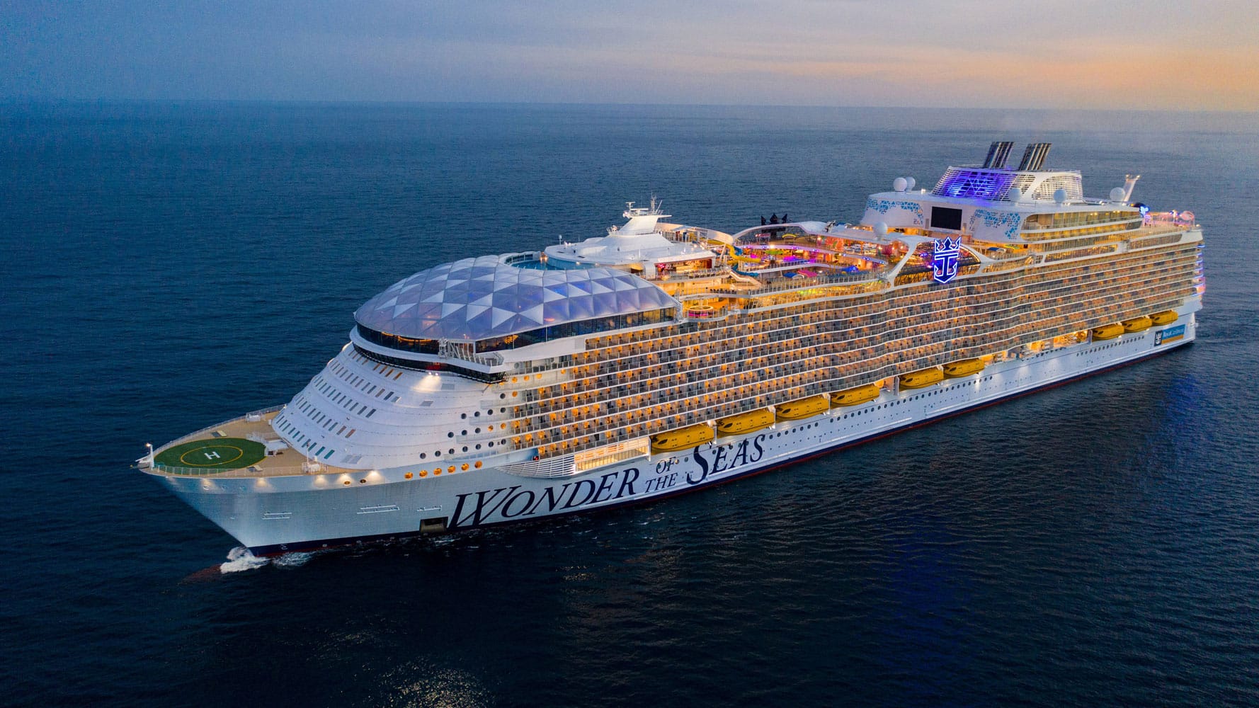 A large white cruise ship, "WONDER OF THE SEAS," sails on dark blue water under a sunset sky.