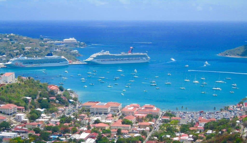An aerial view shows two large cruise ships docked in a turquoise bay filled with many smaller boats.