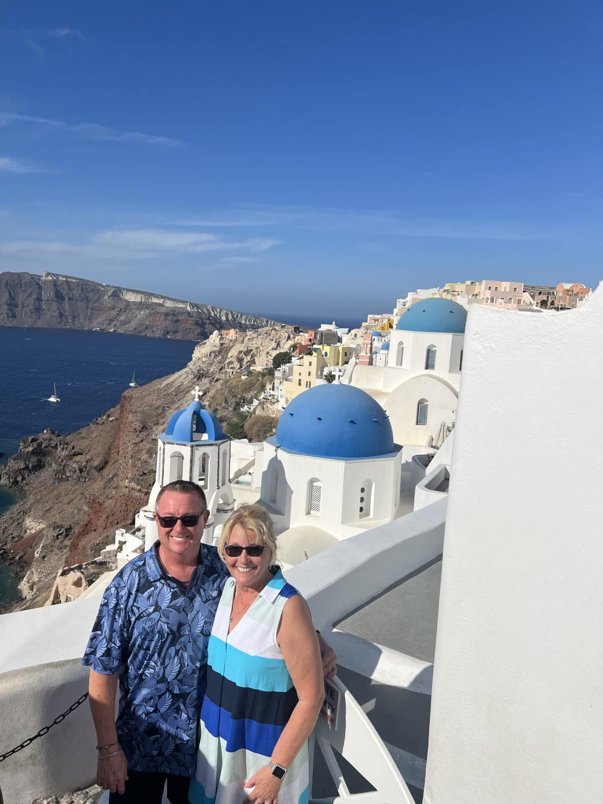 A smiling couple in sunglasses stands on a white balcony overlooking blue-domed churches and a caldera.