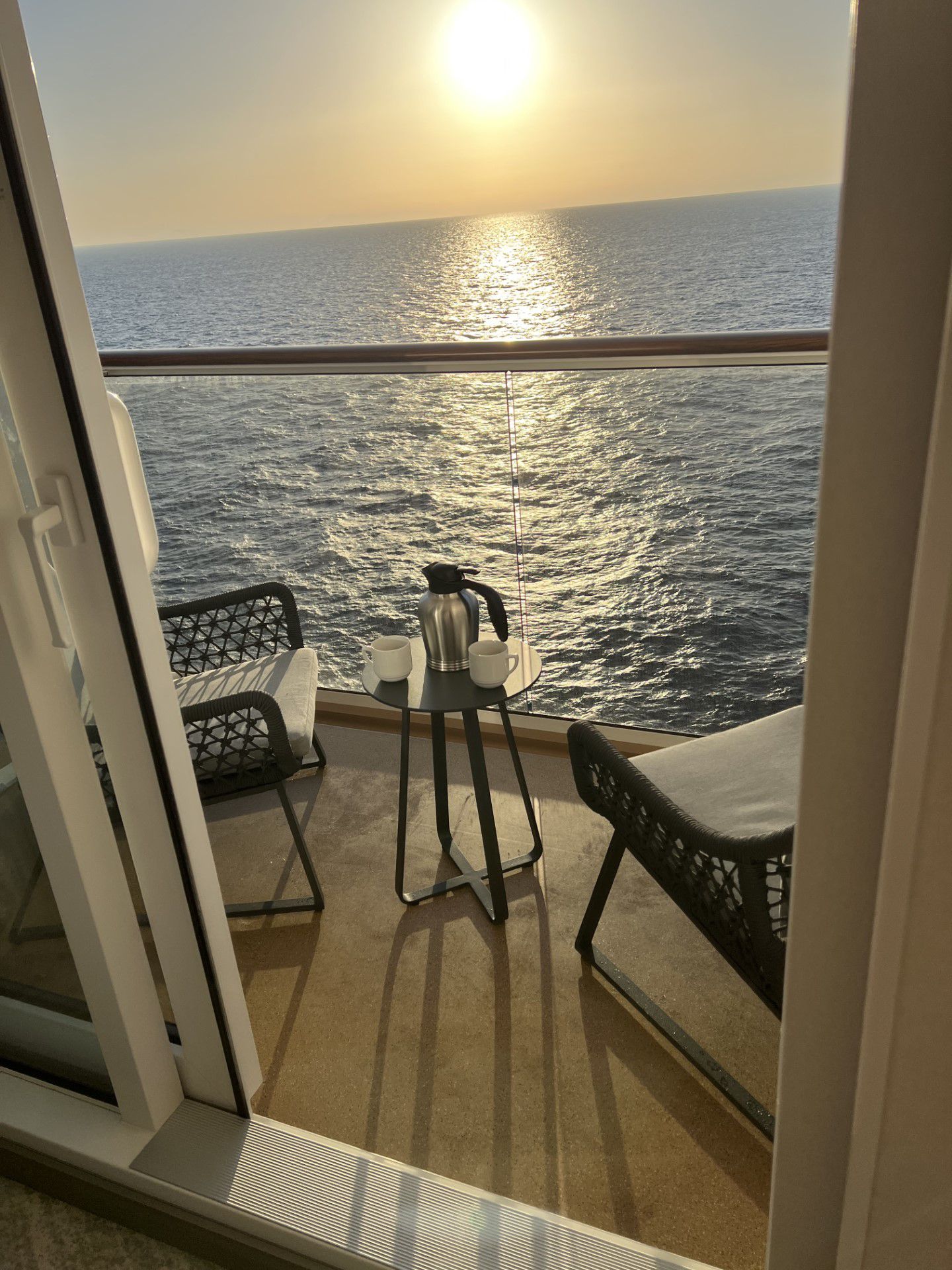 A cruise ship balcony at sunset with a small table, two chairs, a coffee pot, and two mugs.