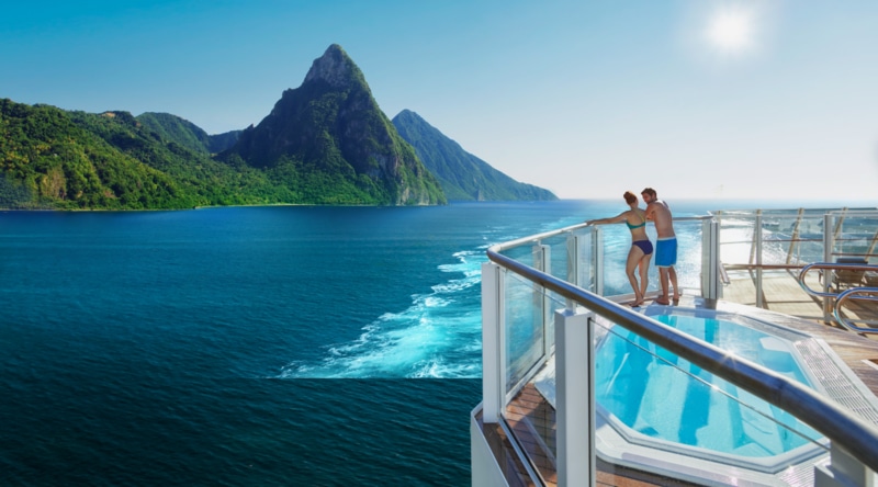 A couple in swimsuits stands on a cruise ship deck overlooking green mountains and blue ocean with a hot tub nearby.